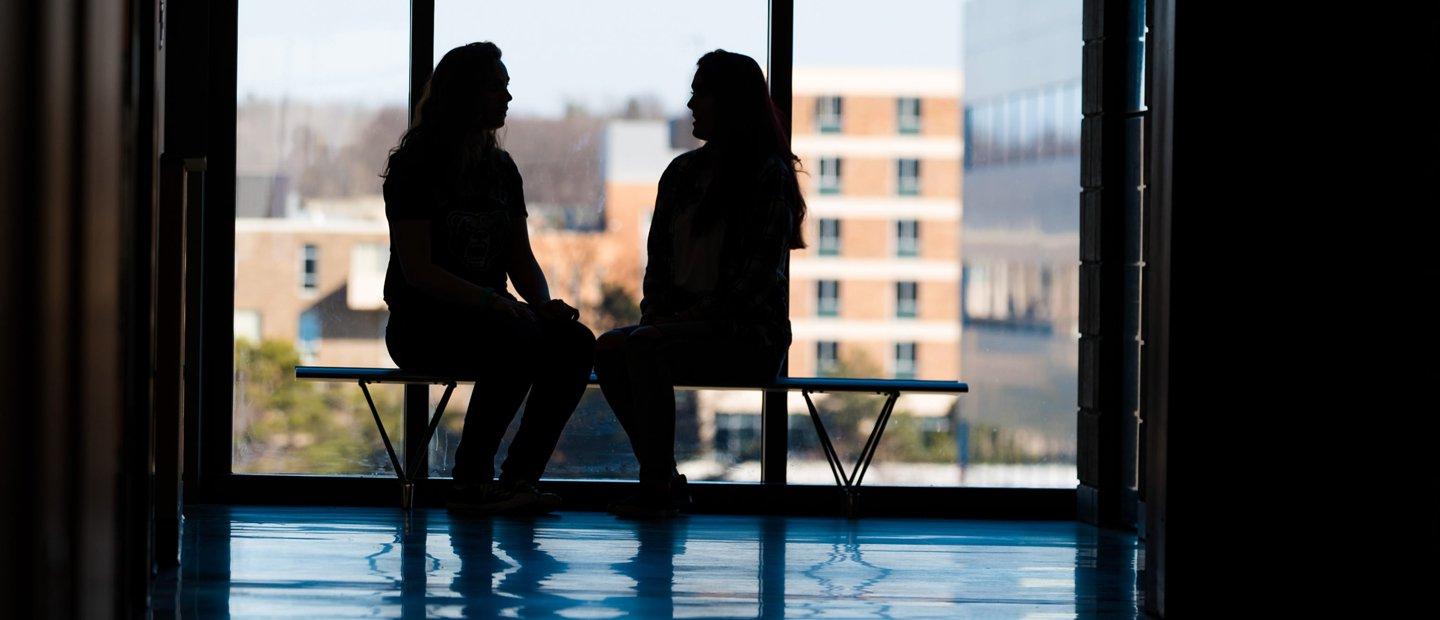 两个人的剪影 seated on a bench in front of a window with O U campus in the background